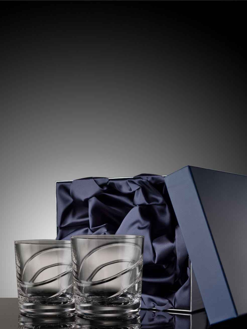 Two clear glass tumblers with a decorative pattern on a dark gray background with a blue satin box.