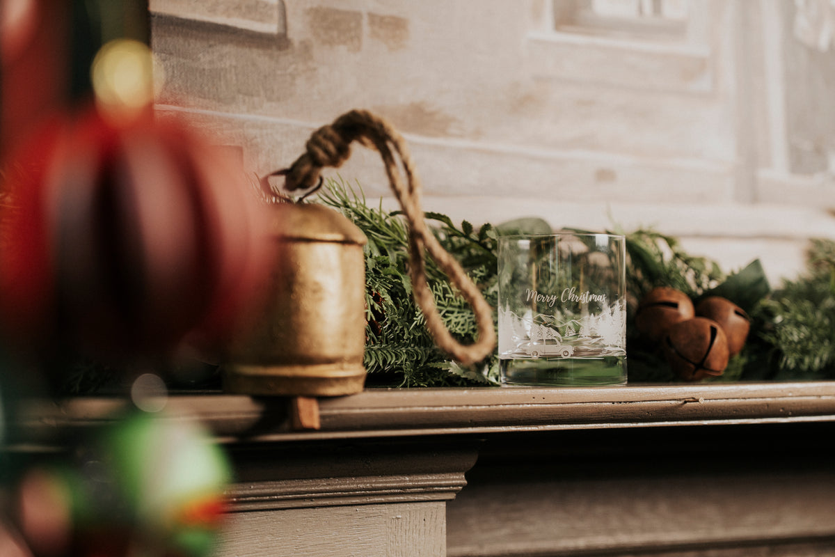 A rustic holiday mantel scene with a brass bell, green garland, a clear glass tumbler, and small wooden ornaments against a softly blurred interior background.