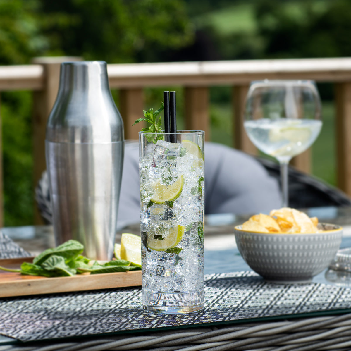 A tall clear glass cocktail tumbler with a black straw and mint garnish, filled with ice and lime slices, set on an outdoor patio next to a stainless steel shaker, a wine glass, and a bowl of chips.