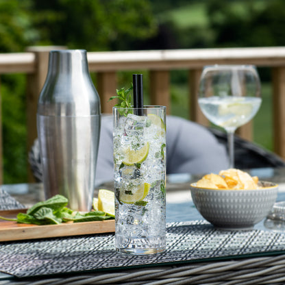 A tall clear glass cocktail tumbler with a black straw and mint garnish, filled with ice and lime slices, set on an outdoor patio next to a stainless steel shaker, a wine glass, and a bowl of chips.